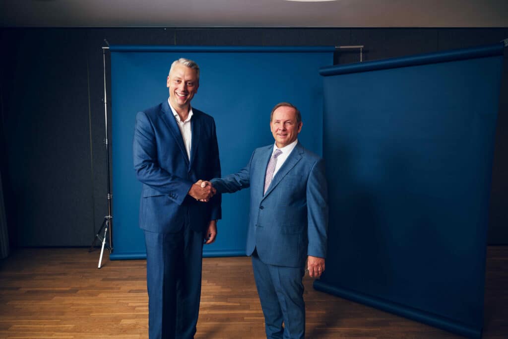 Patrick Oestreich und Dr. Jörg Mosolf schütteln sich die Hände und blicken lächelnd in die Kamera